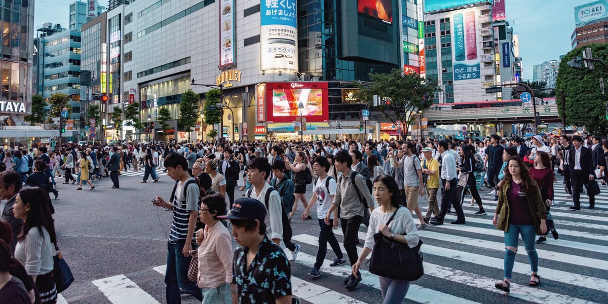 Hundreds of people cross a wide city intersection at dusk beneath illuminated billboards and office towers.