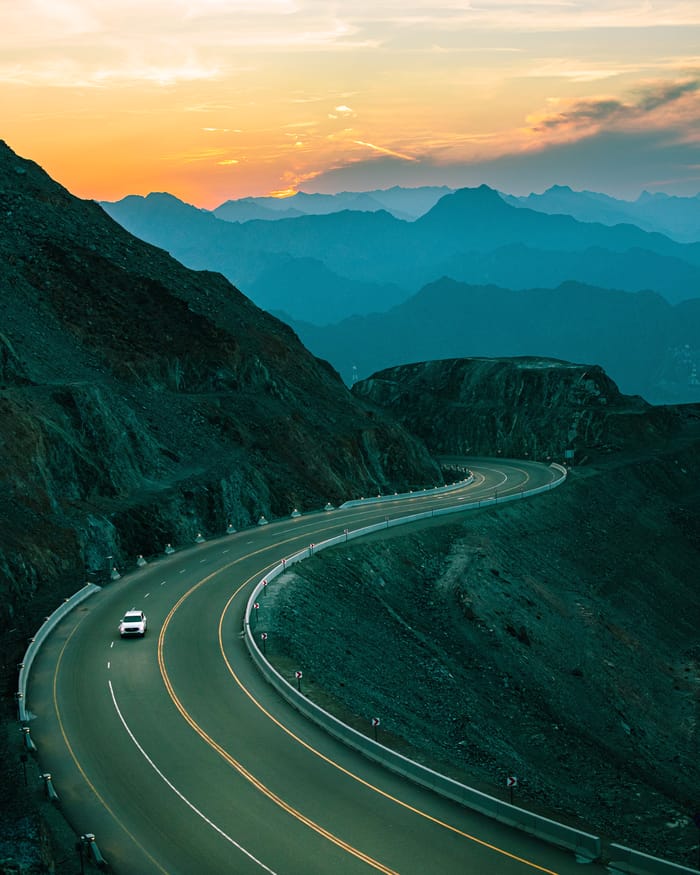 A single car climbs a winding mountain road at dusk, with layered blue ridges and an orange sunset on the horizon.